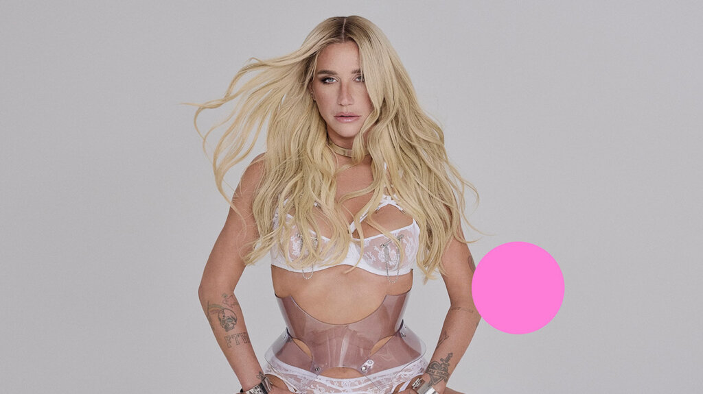 Kesha, a singer with long blonde hair, poses confidently in a white bra against a neutral background.