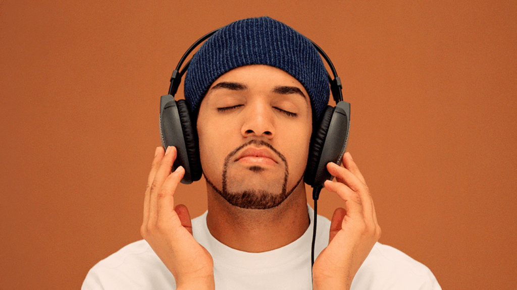 Singer Craig David with a beanie and white t-shirt, listening to music against an orange background.