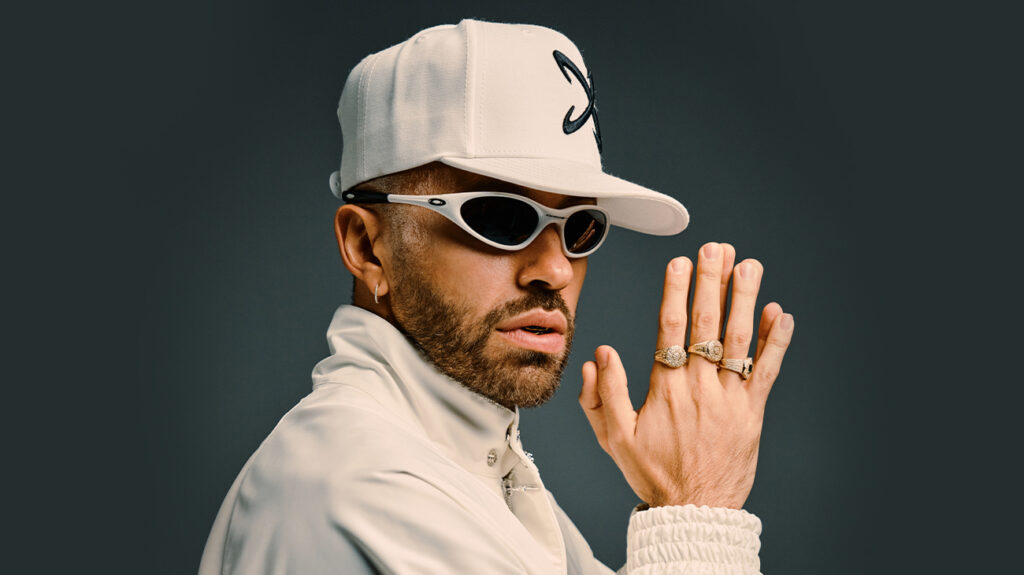 Feid dressed in white and wearing sunglasses poses seriously for the camera.