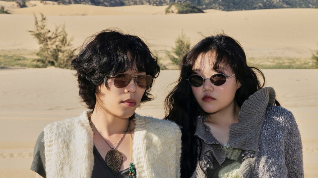Two individuals wearing sunglasses stand together in a vast desert landscape under a clear blue sky.