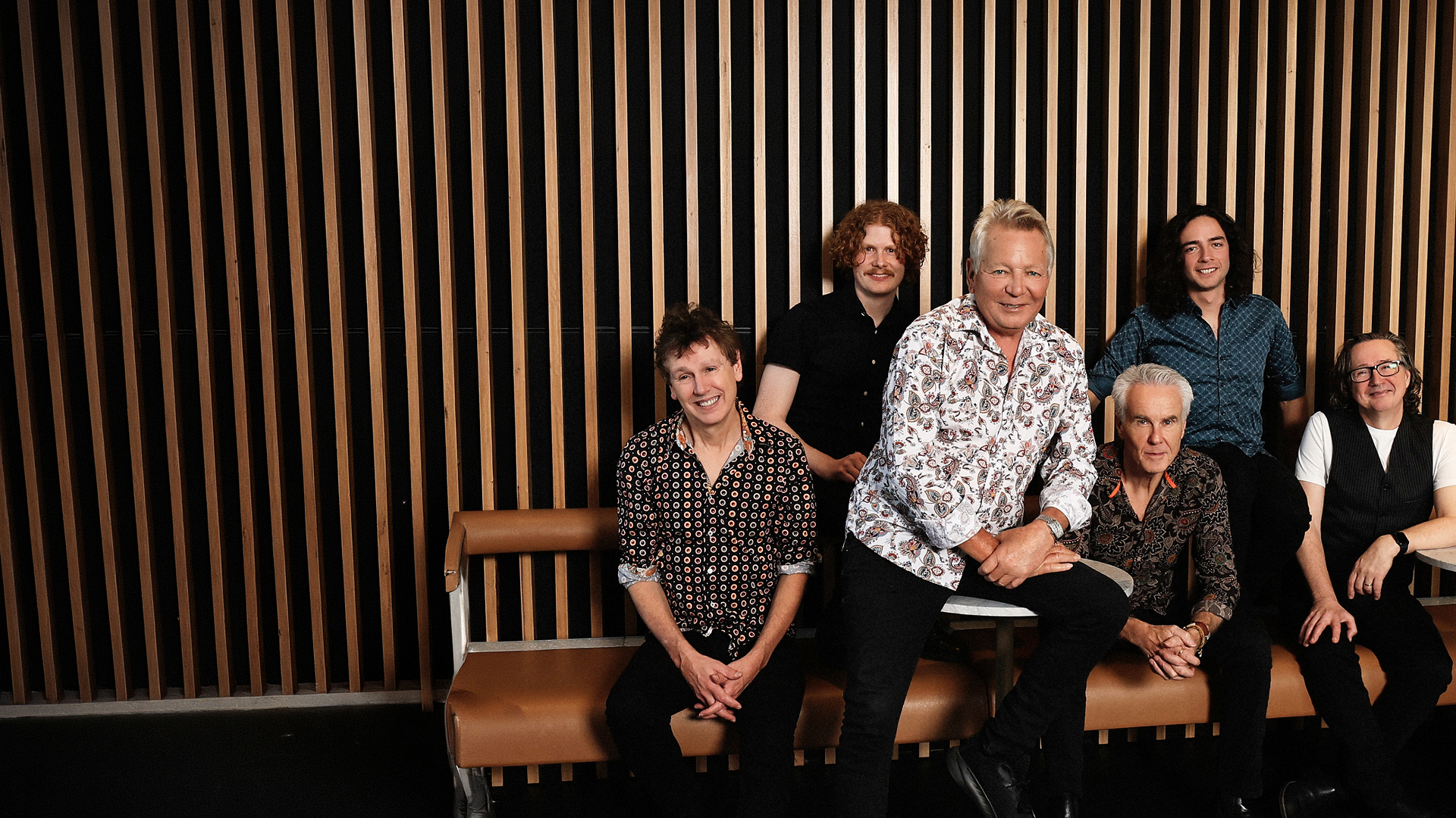 Icehouse band members posing in a room with a wooden wall backdrop.